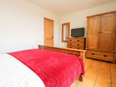 A bedroom with a wooden bed covered by a red blanket a wooden dresser with a TV a mirror a wooden wardrobe and a wooden door at Boslow in St Just