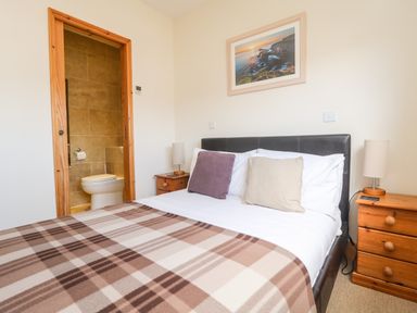 A bedroom with a double bed covered by a plaid blanket and two bedside tables with lamps and a bathroom visible through a wooden door frame at Boslow in St Just