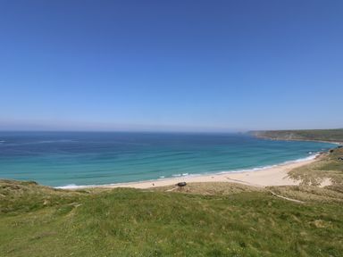 A beach with sand and waves bordered by grassy cliffs under a clear blue sky at Boslow in St Just