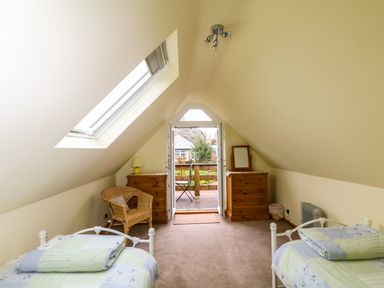 An attic bedroom with two single beds two wooden dressers a wicker chair and a door opening to a balcony at Higher Kernick Stable in Helland near Blisland