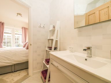 A bathroom with a white sink and ladder shelf with towels and a bedroom with pink curtains visible through the doorway at Higher Kernick Stable Helland near Blisland