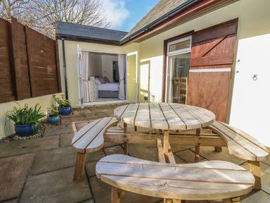 A courtyard with a round wooden picnic table and benches outside a room with open double doors showing a bedroom at Higher Kernick Stable Helland near Blisland