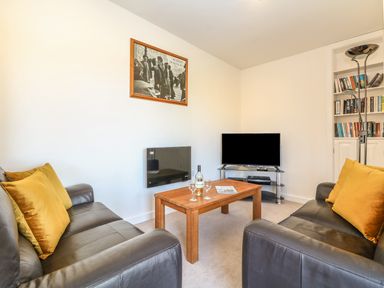 A living room with two black leather sofas with yellow cushions a wooden coffee table with a bottle and glasses a black TV on a stand and a built in bookshelf at Higher Kernick Stable Helland near Blisland