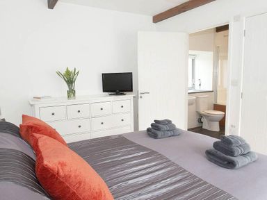A bedroom with a bed with folded towels and orange cushions and a white dresser with a TV and flowers at The Old Dairy in Padstow