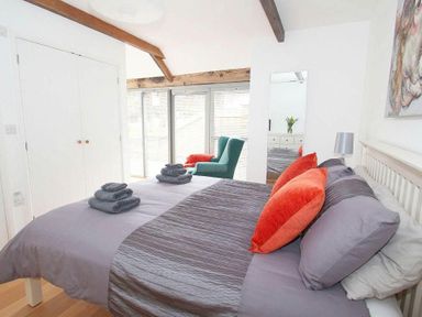 A bedroom with a bed with gray bedding and orange pillows a green armchair a wardrobe and a mirror at The Old Dairy in Padstow