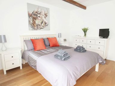 A bedroom with a double bed gray bedding and orange pillows two bedside tables with lamps a dresser with a TV and a vase with flowers and a painting of a bull's head on the wall at The Old Dairy in Padstow