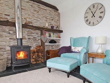 A living room with a lit wood stove fire turquoise chairs and ottomans a wooden side table and a large wall clock at The Old Dairy in Padstow