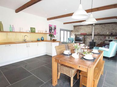 A kitchen and dining area with wooden table and chairs and a living room with teal armchairs and a wood stove at The Old Dairy in Padstow