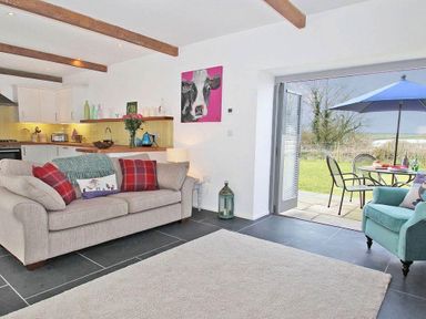 A living area with a beige sofa with red pillows and a blue armchair next to an open door leading to a patio with a blue umbrella at The Old Dairy in Padstow