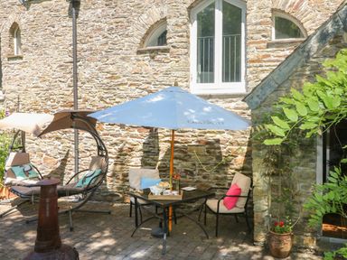 An outdoor patio with a table and umbrella two cushioned chairs and a swing seat against a stone building with arched windows