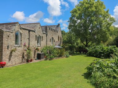 A garden with a large tree and green plants next to a stone building under a blue sky at Hideaway in Watergate Bay