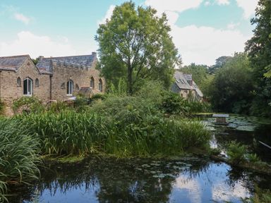 A pond with water plants and stone buildings surrounded by trees at Hideaway in Watergate Bay