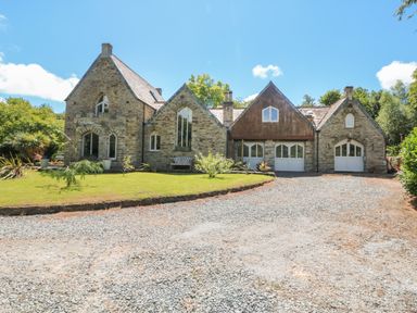 A stone house with multiple gable roofs a gravel driveway and a lawn with plants and trees at Hideaway in Watergate Bay