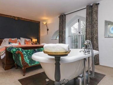 A bedroom with a bed a green upholstered bench and a freestanding bathtub near windows with curtains at Hideaway in Watergate Bay