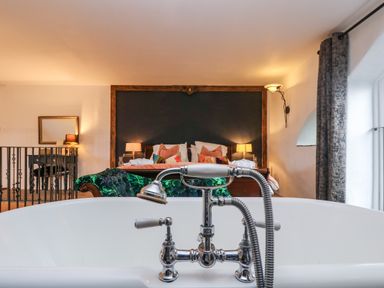 A bathroom with a white bathtub and chrome faucet looking into a bedroom with a wooden bed frame green upholstered chair and lamps at Hideaway in Watergate Bay