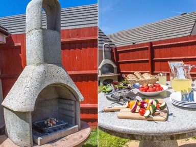 An outdoor stone barbecue grill and a round table with food and drinks in a backyard at Fairways in Woolacombe