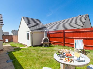 A backyard with a concrete barbecue a table with food and a chair near a red fence at Fairways in Woolacombe