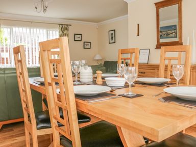 A dining room with a wooden table set with plates glasses and cutlery surrounded by wooden chairs with a green sofa in the background at Fairways in Woolacombe