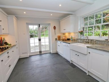 A kitchen with white cabinets and gray tiled floor with a double door and a window showing greenery outside at Mengarth in Cadgwith