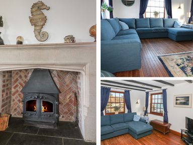 A living room with a stone fireplace with fire a blue sectional sofa wooden floors and windows with blue curtains at Mengarth in Cadgwith