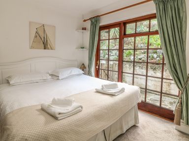A bedroom with a white bed and folded towels on a beige blanket next to large wooden framed windows with green curtains at Mengarth in Cadgwith