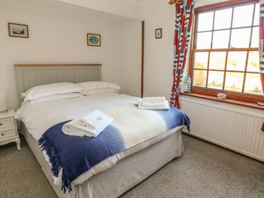 A bedroom with a double bed with white bedding and folded towels on a blue and white throw near a window with red and blue curtains at Mengarth in Cadgwith
