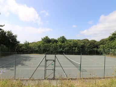 An empty outdoor tennis court surrounded by a chain link fence with trees in the background at Gannet Watch Coverack