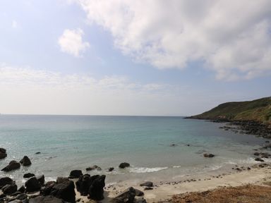A rocky beach with a grassy hill in the background and calm sea under a partly cloudy sky at Gannet Watch in Coverack