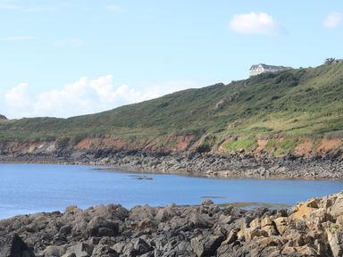 A rocky shoreline with water and a hill with grass and a house on top at Gannet Watch in Coverack