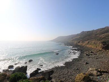 A rocky coastline with waves hitting the shore and hills in the background at Gannet Watch in Coverack