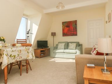 A living room with a dining table a green sofa a beige armchair a wooden TV stand a floor lamp and a telescope at Gannet Watch in Coverack