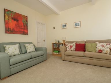 A living room with two sofas, cushions, a small bookshelf with books and a lamp, and paintings on the walls at Gannet Watch in Coverack