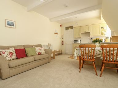 A living room with a beige sofa and colorful cushions a dining table with wooden chairs and a kitchen area at Gannet Watch in Coverack