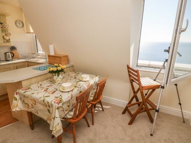 A dining table with two chairs and a floral tablecloth next to a kitchen area and a window with a chair and telescope overlooking the sea at Gannet Watch in Coverack