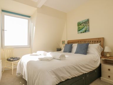 A bedroom with a double bed with white linens and blue pillows near a window and wooden side tables at Gannet Watch Coverack