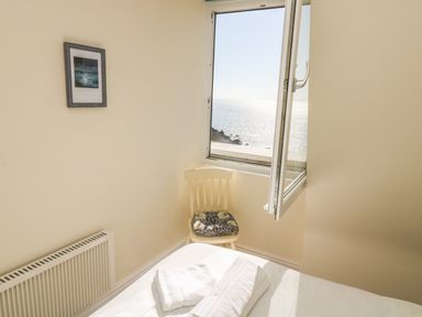 A bedroom corner with an open window showing a sea view a chair with a cushion a radiator and a bed with towels at Gannet Watch in Coverack