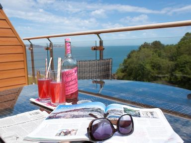 A glass table with two glasses of pink drink a bottle of raspberry lemonade an open magazine and black sunglasses on a balcony with a sea view at Bayview in Brixham