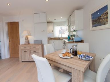 A dining table with breakfast items and white chairs in a kitchen with wood cabinets and a sideboard with a lamp at Bayview in Brixham