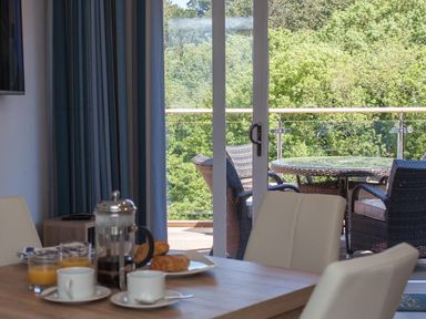 A dining table with coffee cups croissants and orange juice near glass doors opening to a balcony with chairs and a table surrounded by trees at Bayview Brixham
