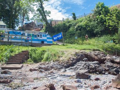 Rocky shoreline with steps leading up to a blue and white building with an open flag at Bayview in Brixham