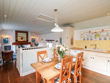 A dining area with a wooden table set with dishes and a vase of flowers next to a kitchen with cream cabinets and yellow tiled backsplash at Trehan in St Ives