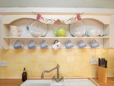 A kitchen shelf with hanging mugs plates teapots and a welcome sign above a sink at Trehan in St Ives
