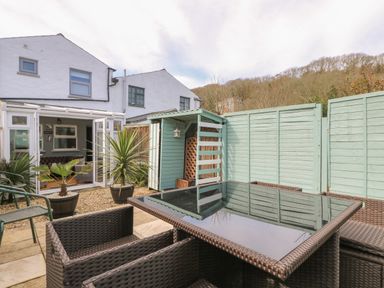A backyard patio with wicker chairs and table two potted plants a small shed and a light blue fence at Trevean in Portreath