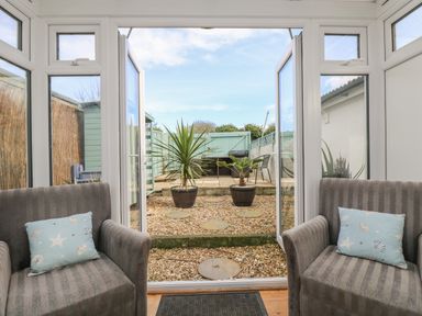 A conservatory with two armchairs and an open glass door leading to a gravel garden with two potted plants and a fence at Trevean in Portreath