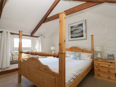 A bedroom with a wooden four-poster bed, two bedside tables with lamps, a framed wall picture, and a window seating area with cushions at Trevean in Portreath