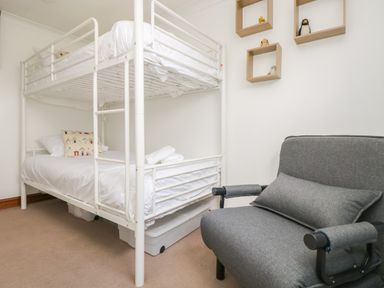 A bedroom with a white metal bunk bed a gray armchair and three wooden wall shelves with small toys at Trevean in Portreath