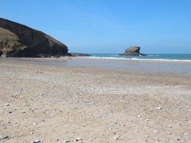 A sandy beach with scattered shells and rocks with a large rock formation on the left and an isolated rock in the sea at Trevean in Portreath
