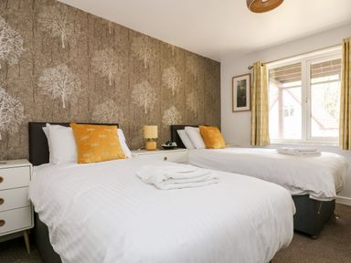 A bedroom with two beds white bedding yellow cushions bedside tables a lamp and wallpaper with tree patterns at Devine Lodge in Callington