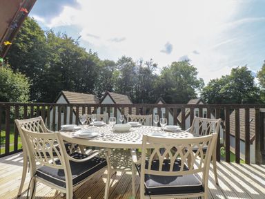 An outdoor dining table with six chairs set with plates and glasses on a wooden deck with houses and trees in the background at Devine Lodge in Callington