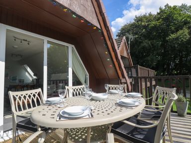 An outdoor dining table with plates and glasses on a wooden deck outside a brown house at Devine Lodge in Callington